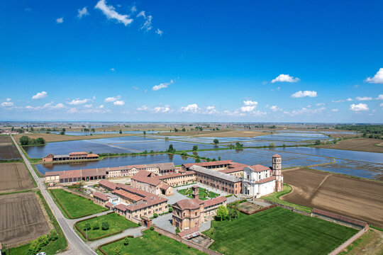 Aerial view of the historic Abbazia di Lucedio complex surrounded by flooded rice fields and rural landscape under a blue sky in Trino, Piemonte, Italy.