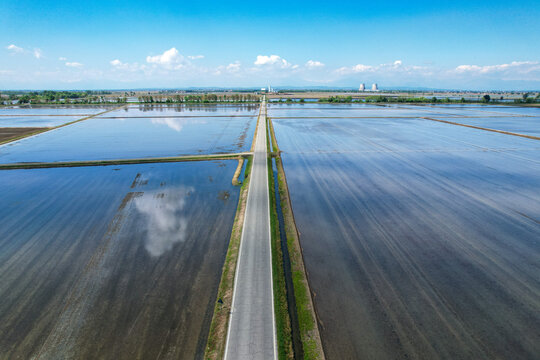 Aerial view of flooded rice fields reflecting the blue sky with a straight road leading towards industrial cooling towers in the distance Trino, Piemonte, Italy.