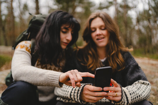 Woman pointing at smart phone held by female friend while spending leisure time in forest