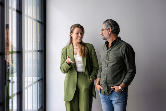 Smiling businesswoman talking with male coworker while standing near wall in office
