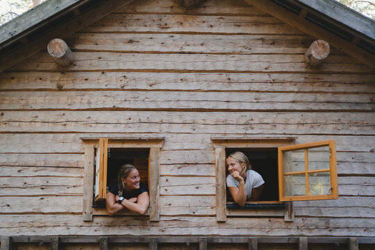 Female friends looking at each other while leaning out from window of log cabin