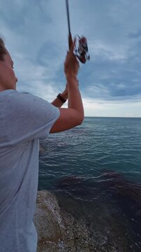 Vertical video. Rear View of Young Woman Casting Spinning Rod Above Tropical Ocean Under Dramatic Cloudy Sky, Slow Motion Recreational Fishing and Coastal Freedom