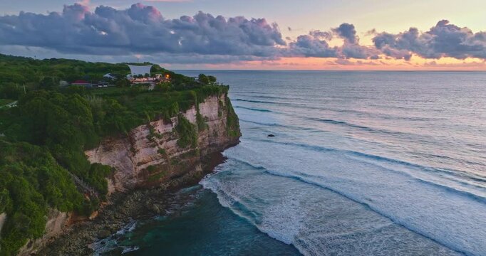 Tropical coastline creates a dramatic cliffside landscape, with green vegetation cascading down to the ocean, and waves breaking under a vibrant sunset sky. Bali travel. Aerial drone flight panorama