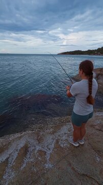 Vertical video. Young Woman Fishing on Rocky Seashore at Cloudy Sunset, back view of female angler casting rod into calm sea under dramatic sky, active leisure and Outdoor Adventure