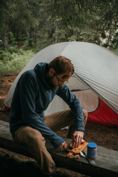 Male explorer cutting apple while sitting on wooden plank near tent in forest