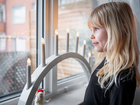 Cute girl with long blond hair standing near electric candles kept on window sill at home