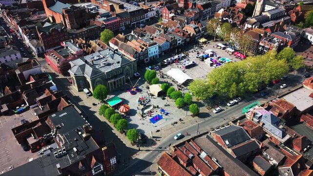 Aerial drone footage pulling back over Salisbury Market Square during a St George&rsquo;s Day event, showing the Guildhall, market stalls, fairground rides, crowds, rooftops, streets,