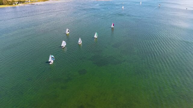 Bird's-eye view of a shallow lake with crystal clear green water and several small sailboats with colorful sails scattered across the surface, revealing ripples and underwater sand patches.
