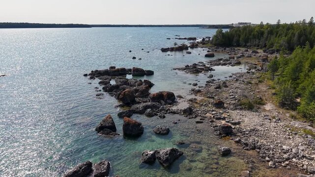 Aerial footage of rocky shoreline along a crystal clear lake in Michigan's Upper Peninsula with scattered boulders and dense evergreen forest on the coast