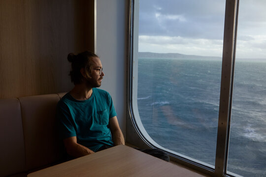 Man wearing eyeglasses looking at sea view through window while traveling in ferry