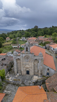 Aerial view of the historic Iglesia de Santa Marina de Augas Santas with its stone facade and red-tiled roof surrounded by village houses and green hills in Augas Santas, Ourense, Spain.