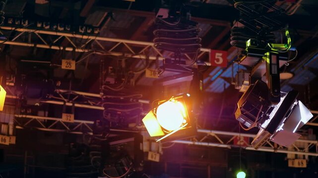 Professional studio stage lighting equipment mounted on ceiling grid illuminating dark film set production area.