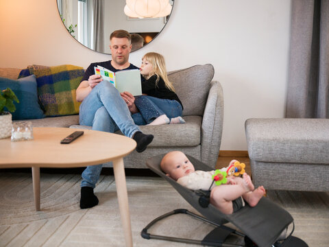 Male toddler lying in carrier while father and daughter reading picture book on sofa at home