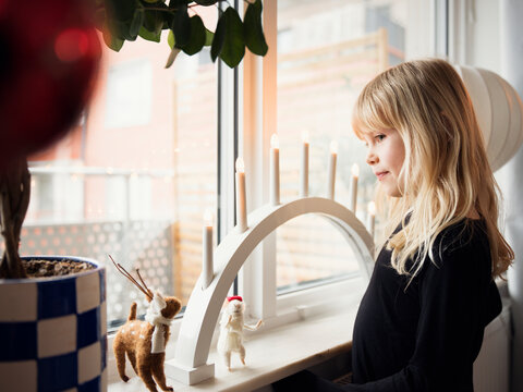 Girl with blond hair standing near needle felted figures kept with electric candle on window sill at home