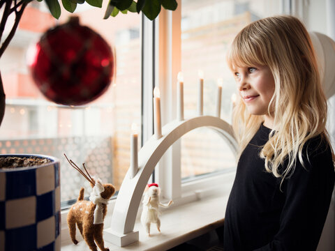 Smiling girl with long blond hair standing near needle felted figures kept with electric candle on window sill at home