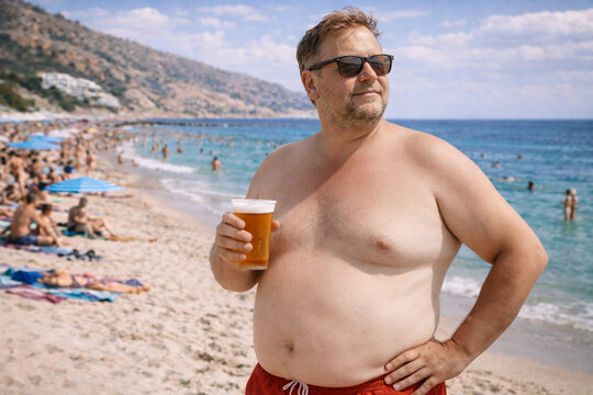 Portrait of a confident overweight man holding a beer on a busy summer beach in Europe.