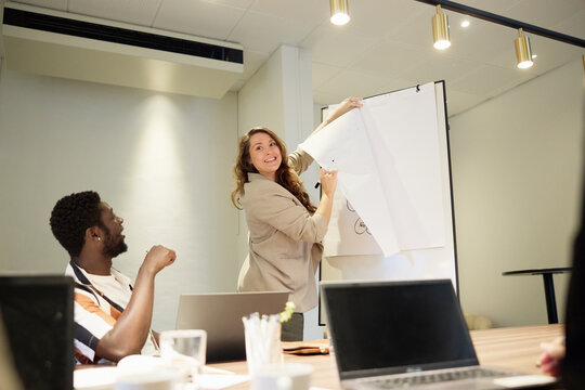 Smiling female manager tearing paper from flipchart while communicating with team in meeting