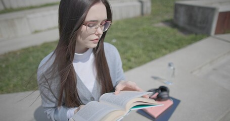 A hand-held shot of a young female student in casual clothes wearing glasses sitting on a concrete bench in an urban park on a sunny summer day and reading a book, flipping through the pages. © Anatoly Repin