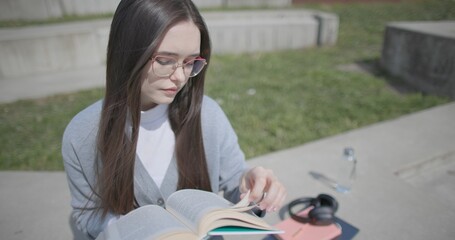 A hand-held shot of a young female student in casual clothes wearing glasses sitting on a concrete bench in an urban park on a sunny summer day and reading a book, flipping through the pages. © Anatoly Repin