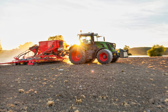 Tractor pulling seed drill across field at sunset