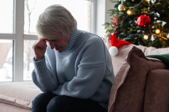 Sad elderly woman sitting on sofa near Christmas tree. Lonely senior feeling depressed during holiday season. Emotional stress, grief, heartache and mental health issue at winter home.