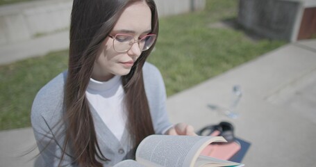 A hand-held shot of a young female student in casual clothes wearing glasses sitting on a concrete bench in an urban park on a sunny summer day and reading a book, flipping through the pages. © Anatoly Repin