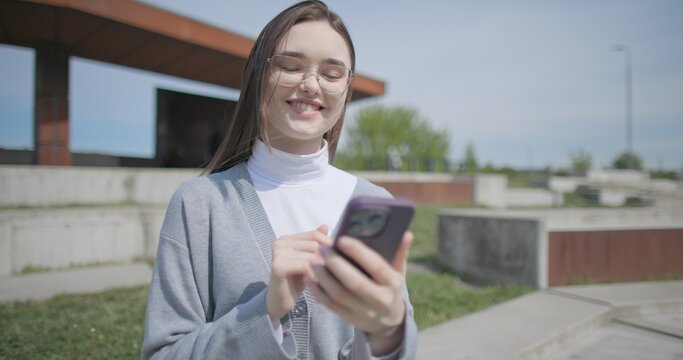 A hand-held shot of a young female student in casual clothes wearing glasses sitting on a concrete bench in an urban park on a sunny summer day using a mobile phone.