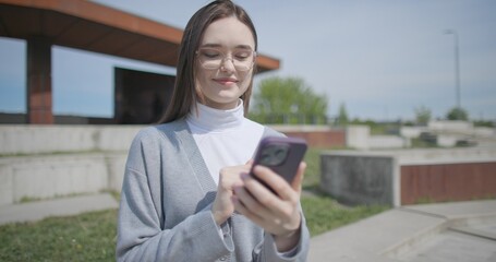 A hand-held shot of a young female student in casual clothes wearing glasses sitting on a concrete bench in an urban park on a sunny summer day using a mobile phone. © Anatoly Repin