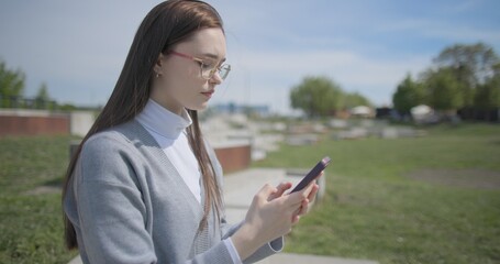 A hand-held shot of a young female student in casual clothes wearing glasses sitting on a concrete bench in an urban park on a sunny summer day using a mobile phone. © Anatoly Repin