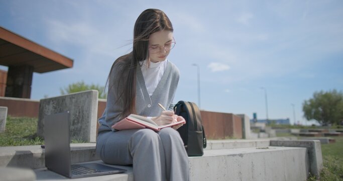 A hand-held portrait of a young female student in casual clothes wearing glasses, sitting on a concrete bench in an urban park, leafing through a notebook and taking notes with a pen.