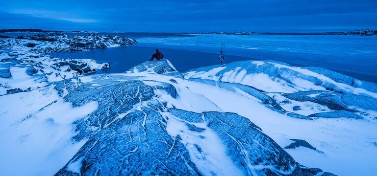 Distant view of person admiring view while sitting on rock amidst frozen lake
