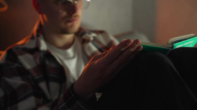 A guy with glasses likes to read a book while sitting on the couch at home in the evening.
Hobbies and leisure, a smart guy flips through pages