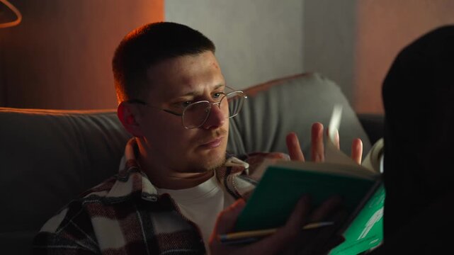 A guy with glasses likes to read a book while sitting on the couch at home in the evening.
Hobbies and leisure, a smart guy flips through pages