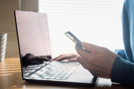 Close-up of man holding phone while sitting with laptop at home