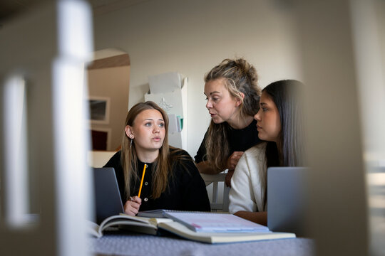 Mother talking with daughter sitting beside friend studying at home
