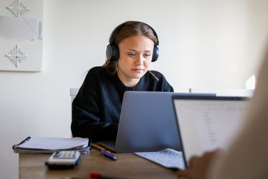 Concentrated teenage girl wearing wireless headphones watching educational lecture on laptop while studying at home