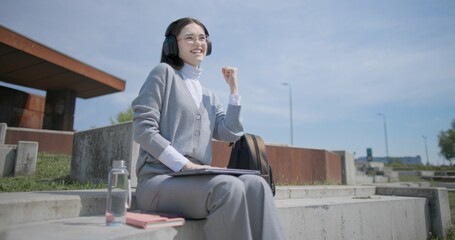 A hand-held shot of a young female student in casual clothes, glasses, and headphones working and typing on a laptop while sitting on a concrete bench in an urban park. © Anatoly Repin