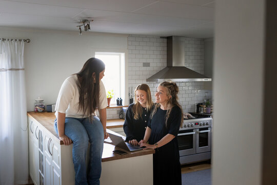 Teenage girl sitting on kitchen island while talking with friend's mother at home