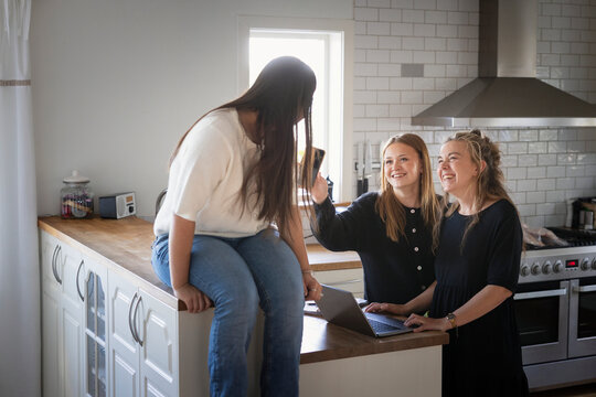 Happy woman standing beside daughter showing smart phone to friend sitting on kitchen island in home