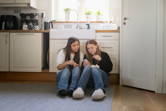 Long haired teenage girl eating popcorn while sitting beside female friend using smart phone at home