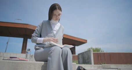 A hand-held shot of a young female student in casual clothes and glasses reading a book while flipping through pages while sitting in an urban park. © Anatoly Repin