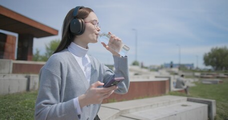 A hand-held shot of a young female student in casual clothes, wearing glasses and headphones, sitting on a concrete bench in an urban park, listening to music and drinking water from a bottle. © Anatoly Repin