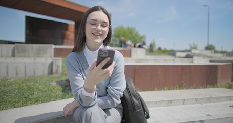 A hand-held portrait of a young female student in casual clothes wearing glasses, sitting on a concrete bench in an urban park and using a phone. © Anatoly Repin