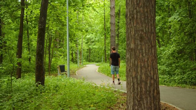 Young man walking alone down a paved path in a lush green park.