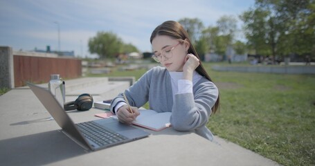 A hand-held shot of a young female student in casual clothes and glasses using a laptop and taking notes with a pen in a notebook in an urban park. © Anatoly Repin