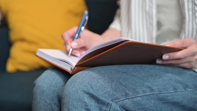 A person writes notes in a notebook while seated on a couch. The individual appears to be focused on their task as they use a pen in a casual indoor setting with soft lighting