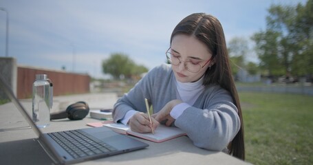 A hand-held shot of a young female student in casual clothes and glasses using a laptop and taking notes with a pen in a notebook in an urban park. © Anatoly Repin