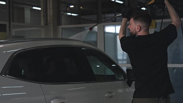 Young man cleaning white crossover SUV with pressure washer at indoor self-service car wash. Professional vehicle washing and detailing concept