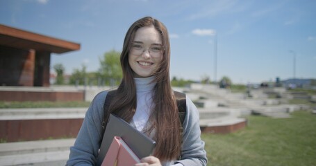 A hand-held portrait of a young female student in casual clothes, wearing glasses and a backpack, holding a notebook and laptop in her hands and posing looking at the camera while standing in a park  © Anatoly Repin