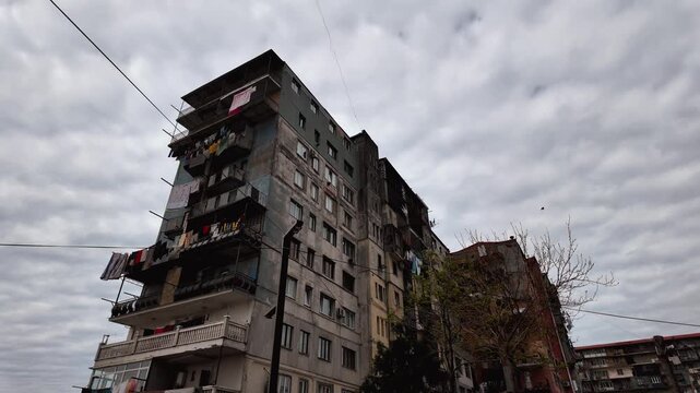 A dirty concrete apartment building under a dramatic cloudy sky, balconies with hanging laundry, dilapidated post-Soviet social housing showing the decay of the city and the gloomy residential life of